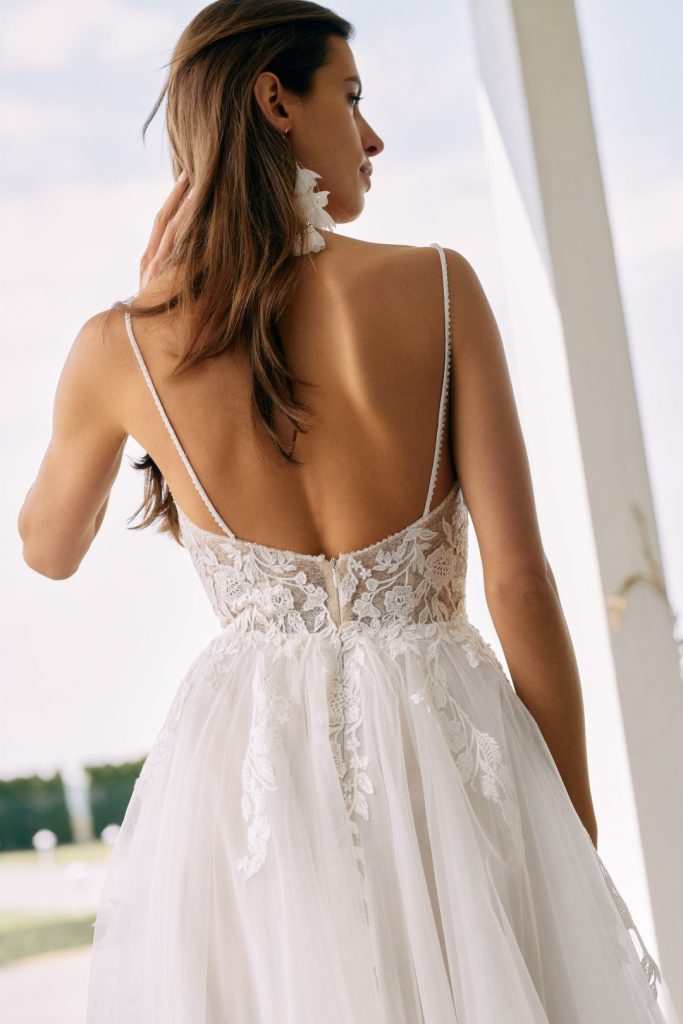 Rückansicht einer Braut, die ein elegantes A-Linien Hochzeitskleid mit tiefem Rückenausschnitt und detailreicher Spitzenverzierung trägt, kombiniert mit auffälligen blütenförmigen Ohrringen.