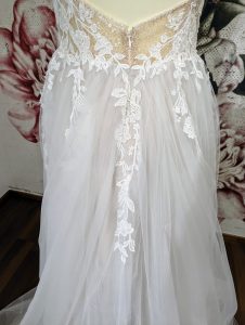 Detailansicht eines Brautkleids, das durch seinen eleganten Spitzenausschnitt und den daran anschließenden fließenden Tüllrock besticht, vor einem Hintergrund mit einem großen floralen Print.
