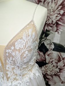 Detailaufnahme eines Brautkleid-Schulterträgers mit zarten Spitzenapplikationen und funkelnden Akzenten, vor einem Hintergrund mit floralem Muster.