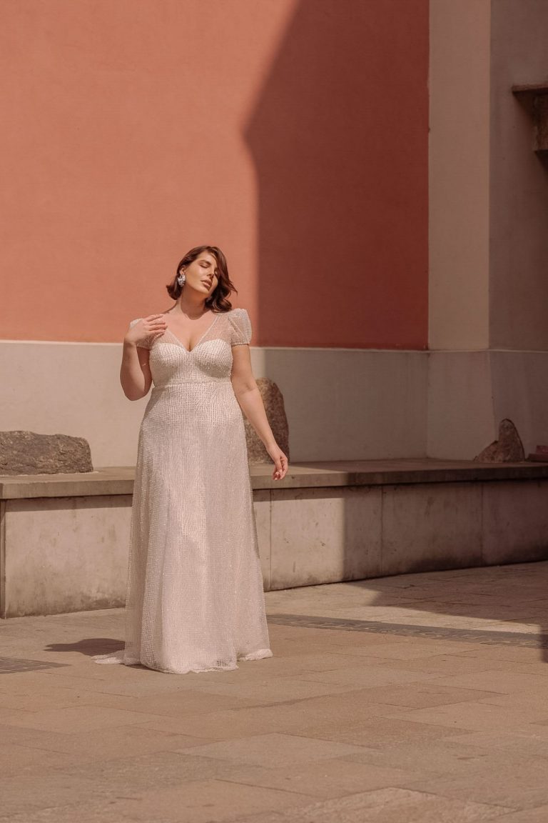 Plus-Size Brautkleid in A-Linie mit funkelnden Perlen und zarten Flügelärmeln vor einer stilvollen, roséfarbenen Wand.