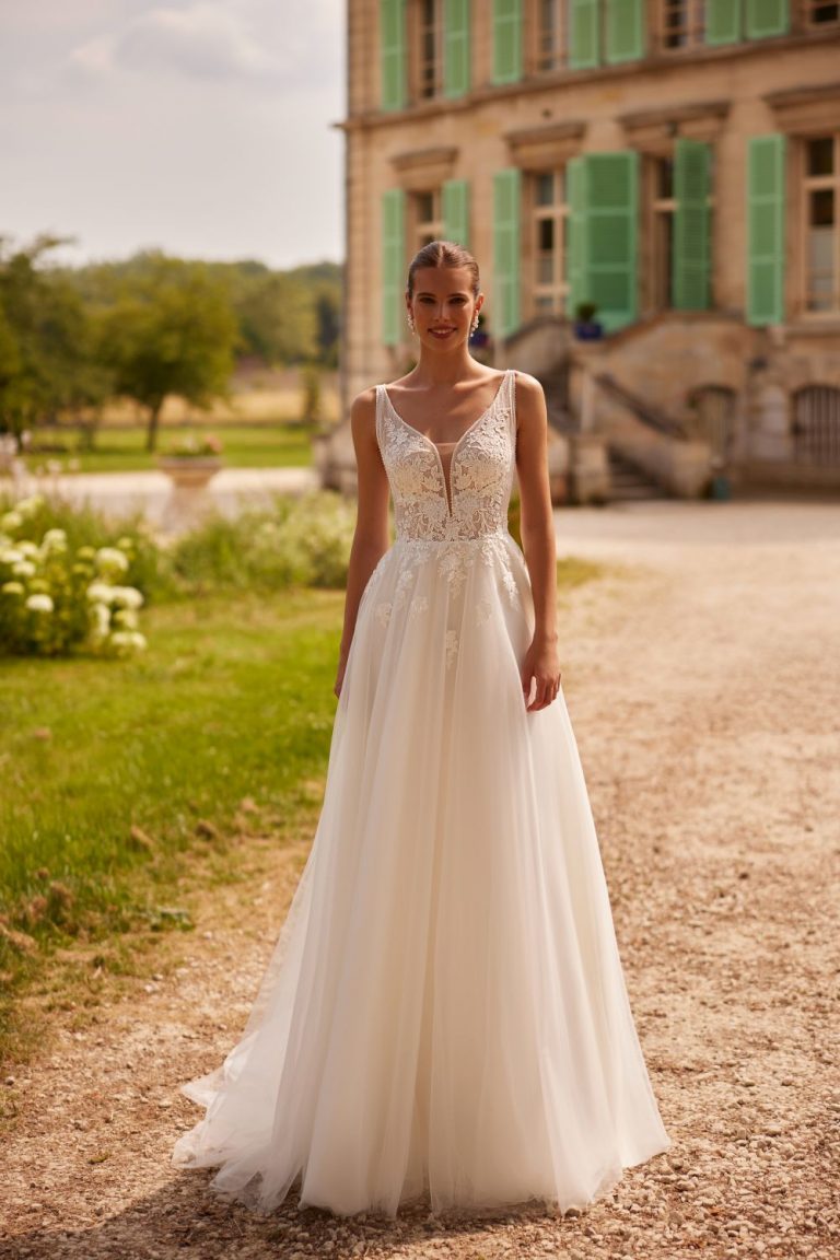 Brautkleid mit floraler Spitze, tiefem V-Ausschnitt und fließendem Tüllrock in der Vorderansicht vor einem historischen Gebäude