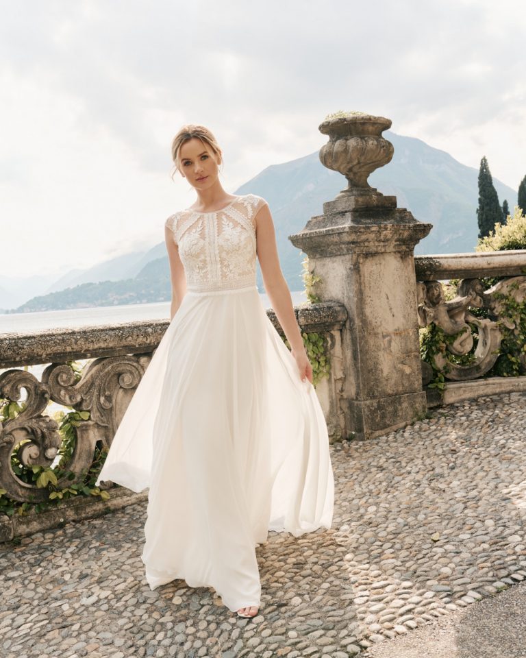 Braut in romantischem Chiffon-Brautkleid mit floraler Spitze und Illusions-Ausschnitt, stehend vor einer malerischen Seelandschaft.
