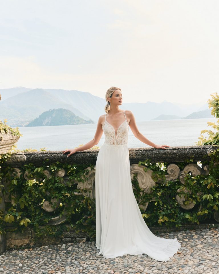 Brautkleid mit tiefem V-Ausschnitt und floraler Spitze, im Vordergrund eine romantische Kulisse mit Blick auf einen See.