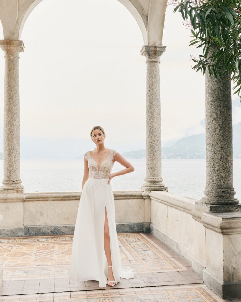 Brautkleid mit tiefem V-Ausschnitt, floralen Spitzenapplikationen und fließendem Chiffonrock mit hohem Schlitz, aufgenommen in einer romantischen Kulisse mit Seeblick.