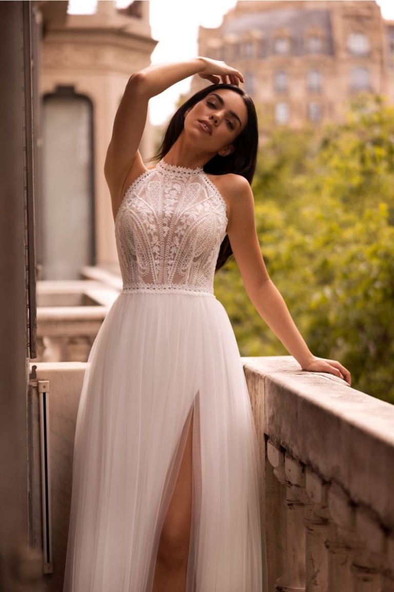 Braut im eleganten Brautkleid mit Spitzenoberteil und Tüllrock, posierend auf einem Balkon mit Blick auf eine romantische Kulisse.