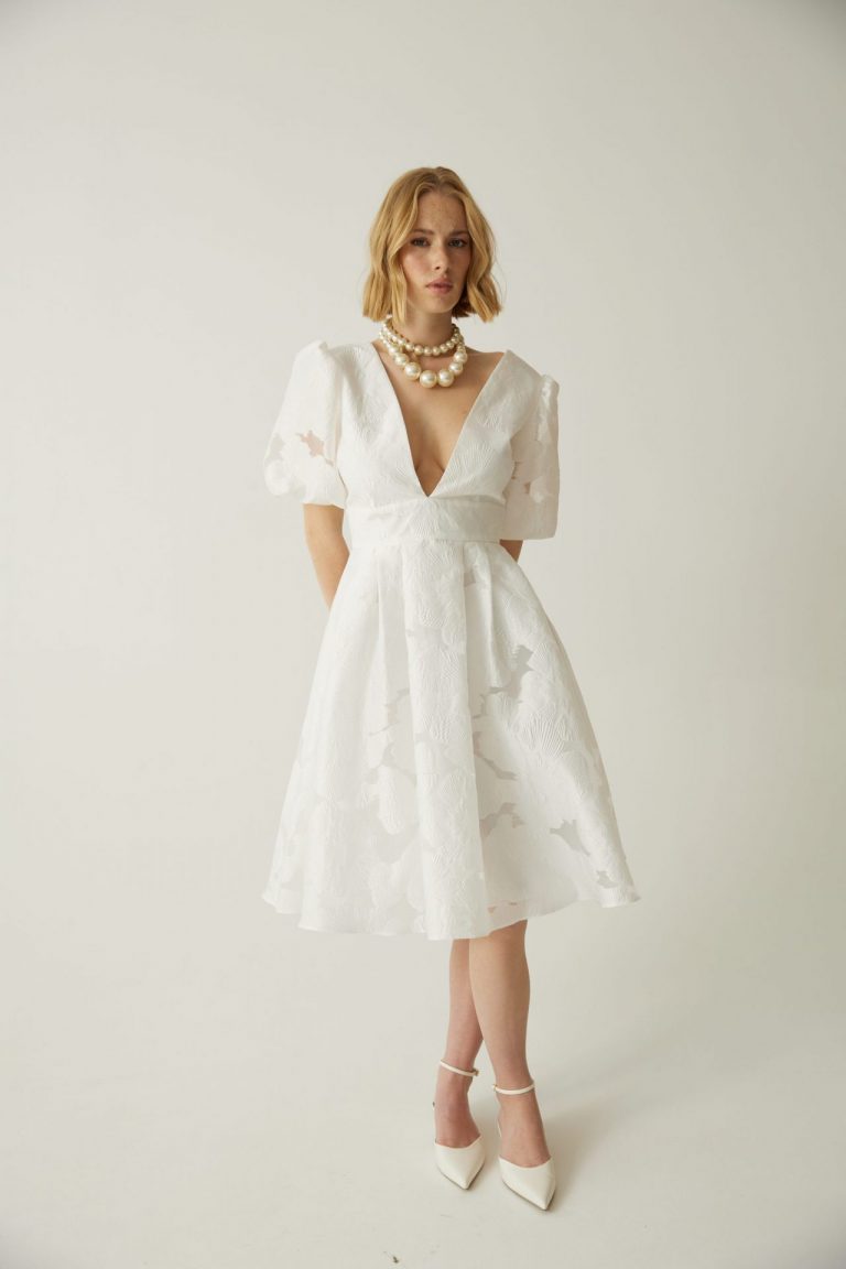 Kurzes Standesamt-Kleid in Weiß mit A-Linien-Schnitt, tiefem V-Ausschnitt, Puffärmeln und floraler Jacquard-Optik, getragen von einer Frau.