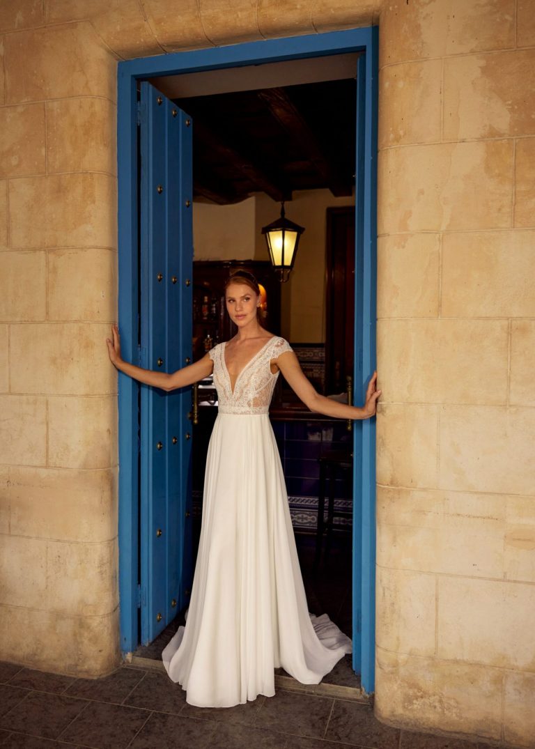 Brautkleid mit tiefem V-Ausschnitt, Spitzenoberteil und fließendem Chiffon-Rock in einem blauen Türrahmen.
