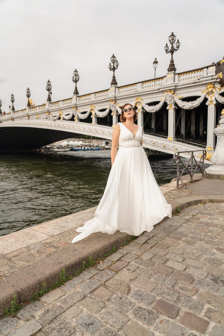 Curvy-Brautkleid mit V-Ausschnitt und floralen Spitzendetails vor einer Brücke in Paris.