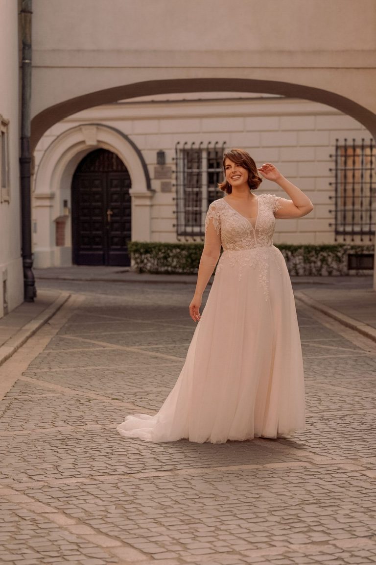 Braut in romantischem Brautkleid mit Spitzenoberteil und Tüllrock, lächelnd in einer gepflasterten Altstadtgasse.