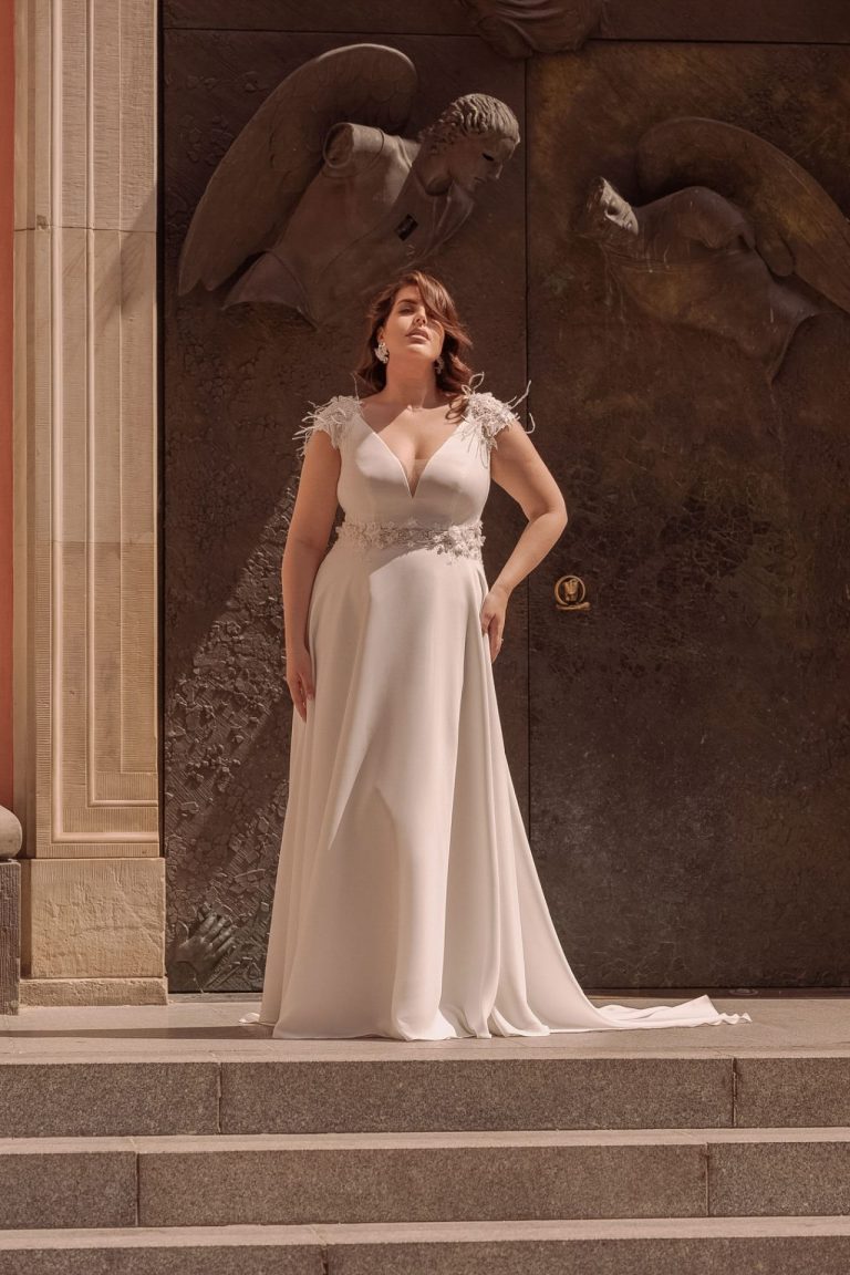 Curvy Brautkleid mit Federn und floraler Spitze, A-Linie, tiefer V-Ausschnitt, romantisch vor antikem Hintergrund.