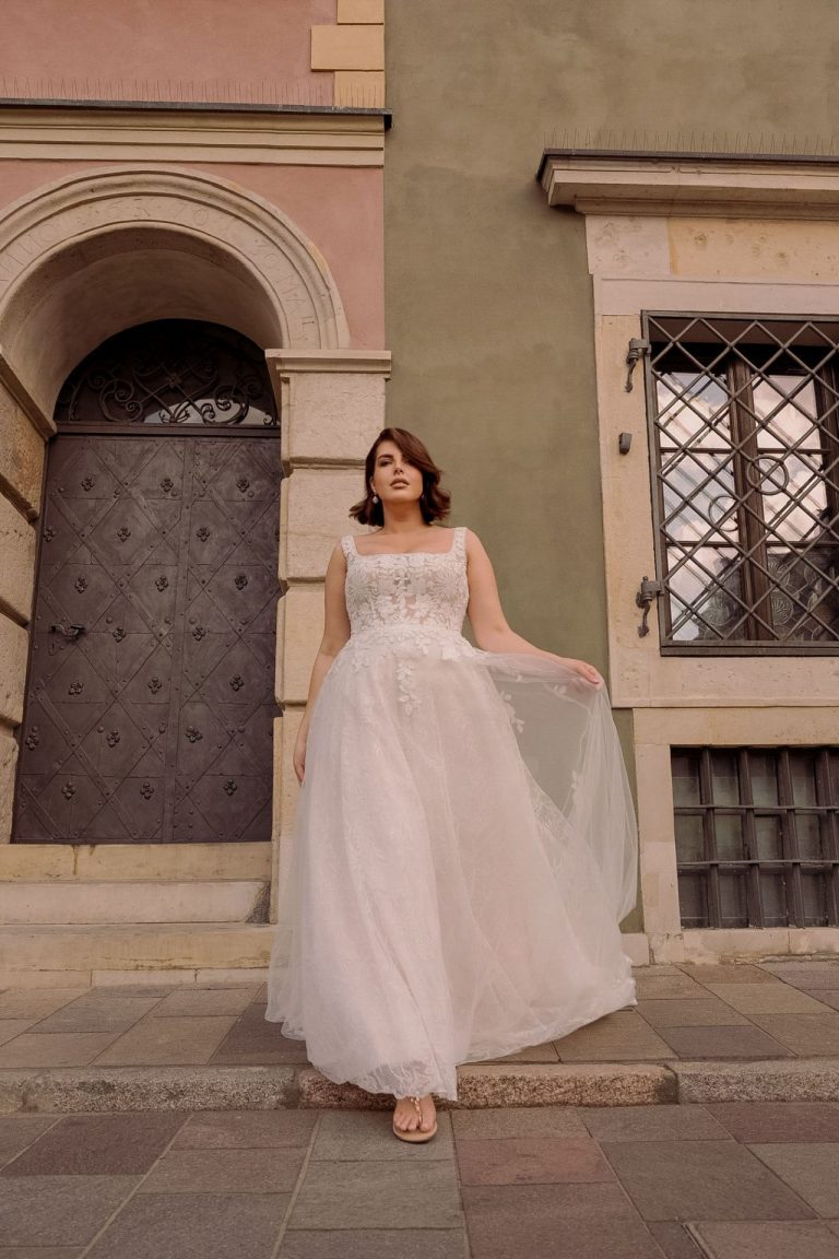 Curvy Braut in einem romantischen Brautkleid mit floraler Spitze vor einer historischen Kulisse